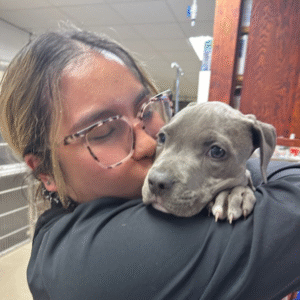 Staff member wearing glasses and a black zip-up jacket kissing a dark-haired dog in a caring moment.